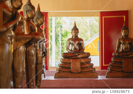 Serene Buddha Statues in a Tranquil Setting with Natural Light Filtering Through Vibrant Red Windows and Lush Greenery in Background 138928673
