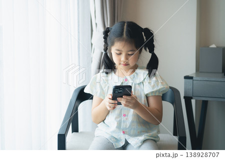 Young girl sitting on chair using smartphone, focused and engaged, indoor setting with natural light, modern home interior, peaceful atmosphere Young girl sitting on chair using smartphone, focused and engaged, indoor setting with natural light, modern home interior, peaceful atmosphere 138928707