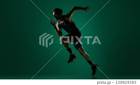 Male athlete performing dynamic sideways jump with extended limbs on dark green background. Male athlete performing dynamic sideways jump with extended limbs on dark green background. 138929165