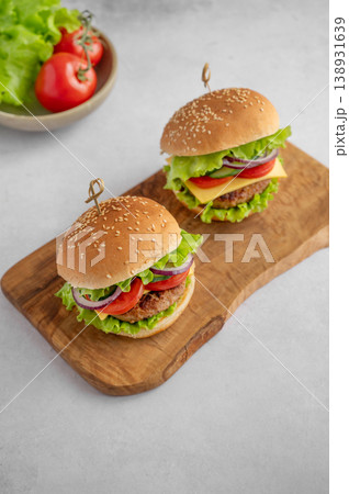 Two beef burgers with tomato, cheese, and lettuce on a cutting board on a light textured background Two beef burgers with tomato, cheese, and lettuce on a cutting board on a light textured background 138931639