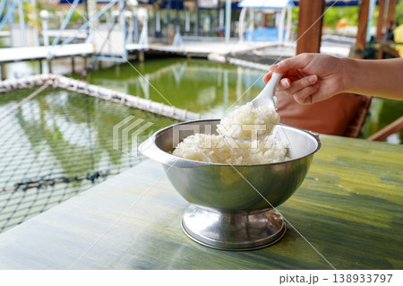 Hand scooping fluffy steamed jasmine rice from a silver serving bowl using a white spoon. The background shows an outdoor waterfront dining area with a green pond and nets. Healthy staple food meal. Hand scooping fluffy steamed jasmine rice from a silver serving bowl using a white spoon. The background shows an outdoor waterfront dining area with a green pond and nets. Healthy staple food meal. 138933797