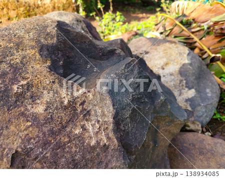 Close up of large rough dark brown boulders resting on dirt ground with blurred green plants, for natural geology and landscape backgrounds. Close up of large rough dark brown boulders resting on dirt ground with blurred green plants, for natural geology and landscape backgrounds. 138934085