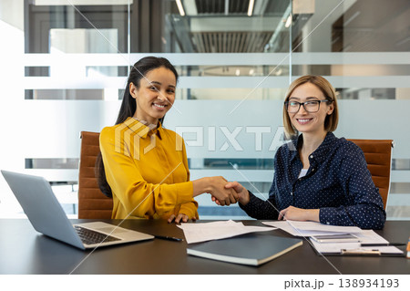 Two smiling multiethnic businesswomen seated at a modern office desk shaking hands after signing documents, celebrating a successful professional agreement and collaborative partnership 138934193