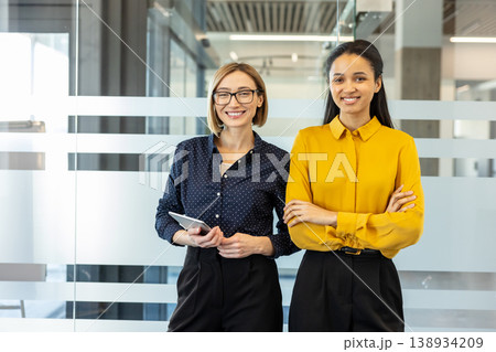 Two smiling professional business women, one holding a tablet and another with crossed arms, standing confidently in a bright contemporary office, illustrating professionalism, teamwork, and success Two smiling professional business women, one holding a tablet and another with crossed arms, standing confidently in a bright contemporary office, illustrating professionalism, teamwork, and success 138934209