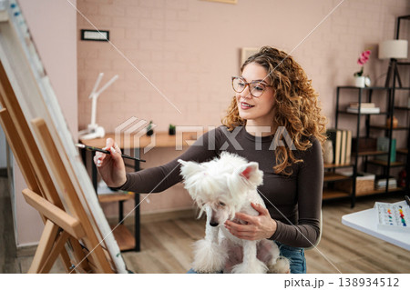 Woman painting while holding her pet dog at home Woman painting while holding her pet dog at home 138934512