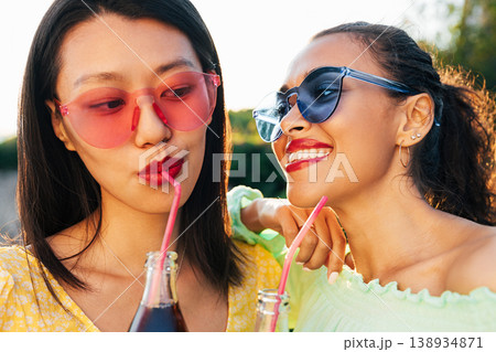 Female friends with drinks. Close-up portrait of two women wearing eyeglasses of different colors while standing outdoors. Female friends with drinks. Close-up portrait of two women wearing eyeglasses of different colors while standing outdoors. 138934871