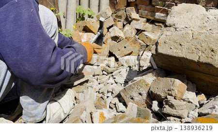 Male hands in protective gloves uses hammer to remove old cement from used bricks during renovation work. The process involves carefully cleaning brick surface to prepare it for reuse in construction. Male hands in protective gloves uses hammer to remove old cement from used bricks during renovation work. The process involves carefully cleaning brick surface to prepare it for reuse in construction. 138936219