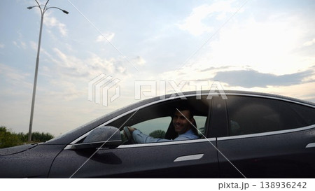 Young businessman driving on electric car in circular path on asphalt road after rain. Happy manager showing grimaces and fooling around while riding on modern auto. Concept of green innovation Young businessman driving on electric car in circular path on asphalt road after rain. Happy manager showing grimaces and fooling around while riding on modern auto. Concept of green innovation 138936242