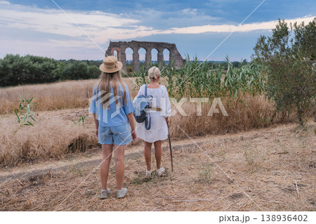 Mother and teenage daughter walking toward ancient Roman aqueduct in Parco degli Acquedotti Rome at sunset through dry grass and natural landscape Mother and teenage daughter walking toward ancient Roman aqueduct in Parco degli Acquedotti Rome at sunset through dry grass and natural landscape 138936402