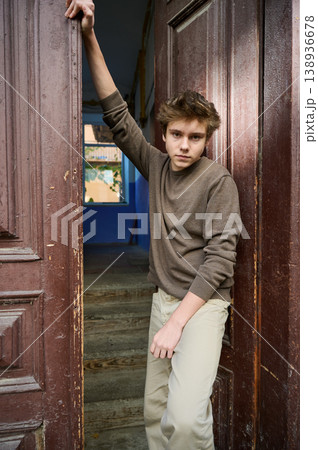 Teen leaning in doorway with gaze, intense eye contact, hand bracing doorframe, textured wood panels, warm interior behind, neutral sweater, confident model energy, cinematic color grading 138936678