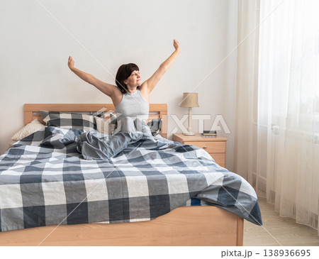 An adult woman feeling refreshed and well-rested, stretching her arms and waking up to natural light in a cozy bedroom. Authentic lifestyle 138936695