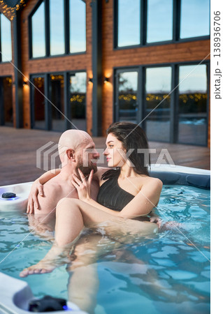 Couple sitting in hot tub looking at each other. Man and woman enjoying intimate spa moment outdoors near modern house, romance, wellness, luxury lifestyle concept 138936706