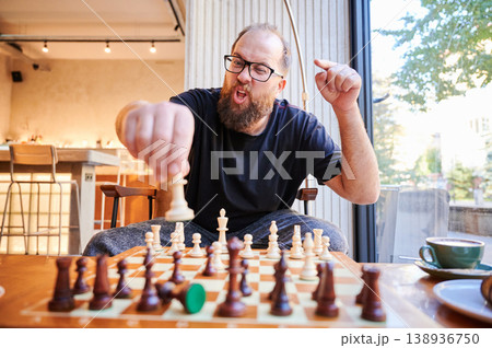 White bearded man concentrates on chess move. Intentional chess player with beard at cafe table during morning. Bearded man carefully plans his next chess move at coffee shop in morning light 138936750