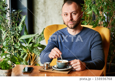 Man stirring coffee and looking at camera in cozy cafe, calm lifestyle scene, relaxed break, modern urban living, everyday routine, quiet moment with drink Man stirring coffee and looking at camera in cozy cafe, calm lifestyle scene, relaxed break, modern urban living, everyday routine, quiet moment with drink 138936768