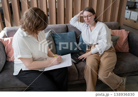 Women sitting on sofa reviewing papers and collaborating in cozy workplace lounge. Collaboration, reviewing, papers, lounge, women, business. Women sitting on sofa reviewing papers and collaborating in cozy workplace lounge. Collaboration, reviewing, papers, lounge, women, business. 138936830