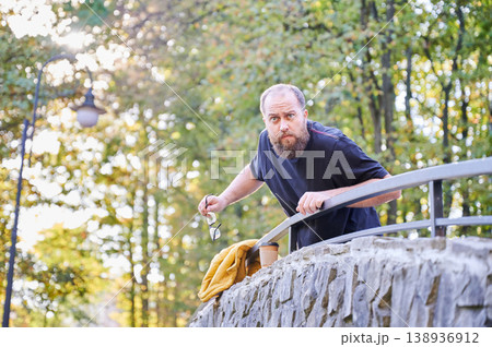 Bearded man searching for keys on stone bridge, peering over railing, holding small keyring, yellow jacket draped over wall, concerned expression, late afternoon light through trees 138936912