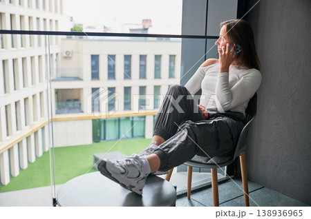 Female resident makes phone call on balcony with city view in contemporary housing complex. Ideal for real estate promotion, connectivity, home lifestyle, and modern living concepts 138936965