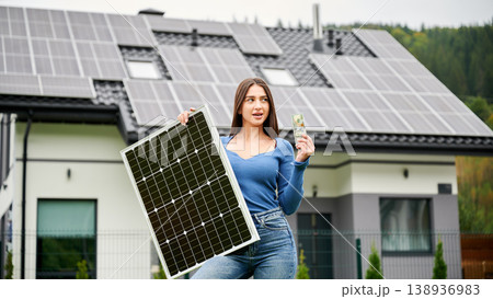 Woman holding solar panel and showing money near house with rooftop panels. Concept of renewable energy savings, eco friendly lifestyle, sustainable home 138936983