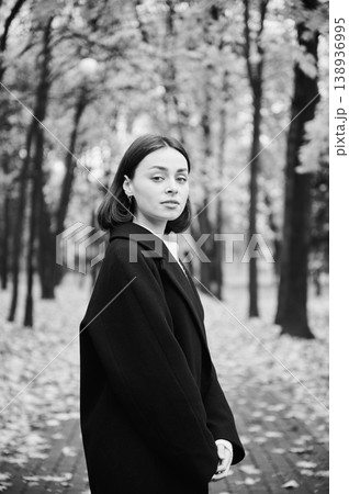 Young woman posing in black coat on park path in monochrome autumn portrait, looking at camera among blurred trees. Ideal for fashion, beauty, timeless style, and advertising Young woman posing in black coat on park path in monochrome autumn portrait, looking at camera among blurred trees. Ideal for fashion, beauty, timeless style, and advertising 138936995