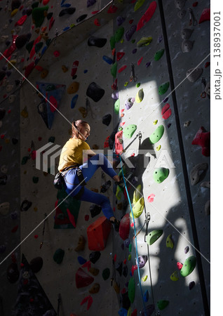 Female climber ascends artificial rock wall with strong shadow and spotlight in sports gym. Great for determination, endurance, motivation, recreation or indoor sport concepts Female climber ascends artificial rock wall with strong shadow and spotlight in sports gym. Great for determination, endurance, motivation, recreation or indoor sport concepts 138937001
