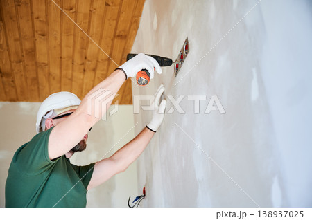 Electrician installing wall outlet with drill and securing electrical system components. Worker in helmet performing interior finishing, symbol of home improvement and modern housing development Electrician installing wall outlet with drill and securing electrical system components. Worker in helmet performing interior finishing, symbol of home improvement and modern housing development 138937025