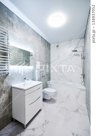 Contemporary bathroom interior with bathtub, suspended toilet and sink cabinet. Refined washroom with cool gray stone texture, white marble wall panels, backlit mirror and towel radiator Contemporary bathroom interior with bathtub, suspended toilet and sink cabinet. Refined washroom with cool gray stone texture, white marble wall panels, backlit mirror and towel radiator 138937052