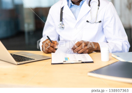 Medical professional in white coat with stethoscope reviews patient files and health records while working on a laptop at a wooden desk, managing documentation and care planning 138937363