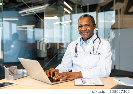 Mature medical professional, a smiling male doctor, working on a laptop at his desk in a modern healthcare office, providing telehealth and digital patient care 138937366