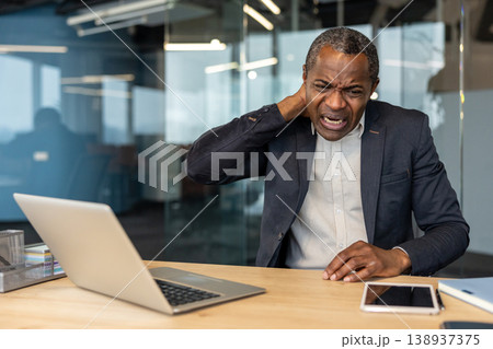 Mature man working on a laptop experiencing discomfort and strain in his neck, holding it with his hand while feeling pain from poor posture or prolonged computer use in an office setting 138937375