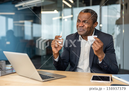 Businessman holding a nasal spray and tissue, experiencing discomfort and allergy symptoms, dealing with a cold or flu while working at his office desk Businessman holding a nasal spray and tissue, experiencing discomfort and allergy symptoms, dealing with a cold or flu while working at his office desk 138937379