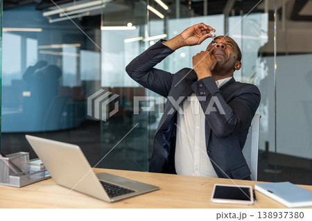 Mature man sitting at a desk in a contemporary office, carefully applying eye drops to relieve discomfort and addressing eye care needs while working on a laptop Mature man sitting at a desk in a contemporary office, carefully applying eye drops to relieve discomfort and addressing eye care needs while working on a laptop 138937380