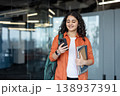 Young indian female student carrying a backpack and notebooks, smiling while looking at her mobile phone, walking through a modern university building 138937391