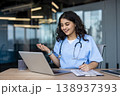 Young female healthcare provider wearing scrubs and stethoscope sitting at desk, having a virtual meeting or teleconsultation with a patient, writing notes while communicating online 138937393