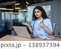 Young indian female doctor wearing scrubs and a stethoscope, showing confusion and concern while having a video call on a laptop in a modern office, expressing frustration 138937394
