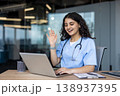 Young female healthcare professional smiling and waving at a laptop screen, engaging in a virtual consultation or remote patient care session from an office environment 138937395