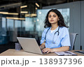 Young female medical professional wearing scrubs and a stethoscope, sitting at a desk and typing on a laptop, representing healthcare innovation and digital medicine 138937396