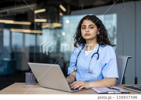 Young female medical professional wearing scrubs and a stethoscope, sitting at a desk and typing on a laptop, representing healthcare innovation and digital medicine 138937396