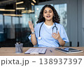Young indian female doctor wearing scrubs and a stethoscope, gesturing while engaging in a professional online remote medical consultation from a modern office environment 138937397