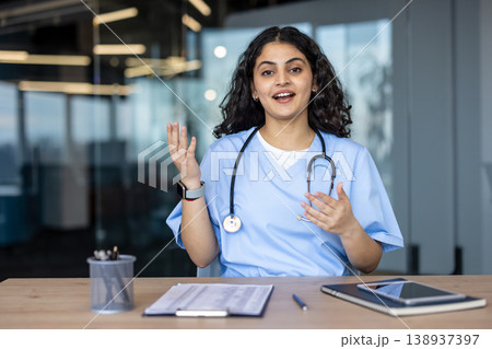 Young indian female doctor wearing scrubs and a stethoscope, gesturing while engaging in a professional online remote medical consultation from a modern office environment 138937397