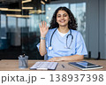 Young female healthcare professional wearing scrubs and stethoscope, sitting at a desk in a modern office, smiling, looking at the camera, and waving her hand, embodying welcome and friendly service 138937398