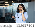 Female medical professional in scrubs and a stethoscope talking on a mobile phone, actively communicating while gesturing, standing in a modern clinic or hospital setting 138937401