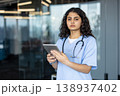 Young female medical professional in scrubs and stethoscope standing while holding a digital tablet, representing modern healthcare, medical technology, and dedicated patient care 138937402