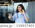 Young woman nurse or doctor wearing scrubs and stethoscope, engaging in a virtual consultation with a patient via video call on a digital tablet, providing telehealth services 138937403