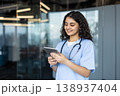 Young female nurse in light-blue scrubs and stethoscope smiling while using a tablet for patient records and telehealth in a modern clinic, showcasing digital healthcare integration 138937404