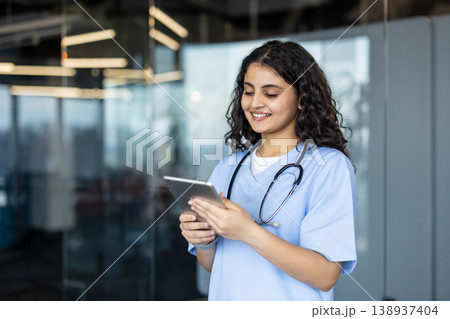 Young female nurse in light-blue scrubs and stethoscope smiling while using a tablet for patient records and telehealth in a modern clinic, showcasing digital healthcare integration 138937404