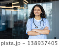 Young female medical professional with a smiling expression, wearing scrubs and a stethoscope, standing confidently with arms crossed in a modern hospital or clinic environment 138937405