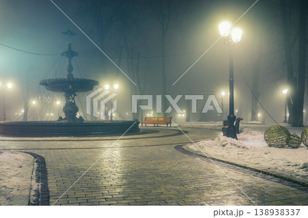 Big fontaine and alley of a night winter park in a fog. Footpath in a fabulous winter city park at night in fog with benches and patterns. Beautiful foggy evening in the Mariinsky Park. Kyiv, Ukraine. 138938337