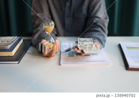 Time and money concept. Businessman holding cash and sand clock imply growth of income, saving or return investment through time. Time and money concept. Businessman holding cash and sand clock imply growth of income, saving or return investment through time. 138940702