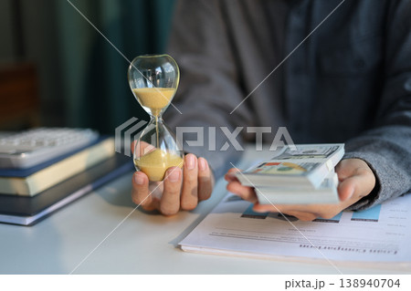 Time and money concept. Businessman holding cash and sand clock imply growth of income, saving or return investment through time. Time and money concept. Businessman holding cash and sand clock imply growth of income, saving or return investment through time. 138940704