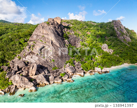 Imposing rocky cliffs rise above lush greenery, with turquoise waters at the base. Seychelles, La Digue. Imposing rocky cliffs rise above lush greenery, with turquoise waters at the base. Seychelles, La Digue. 138941397